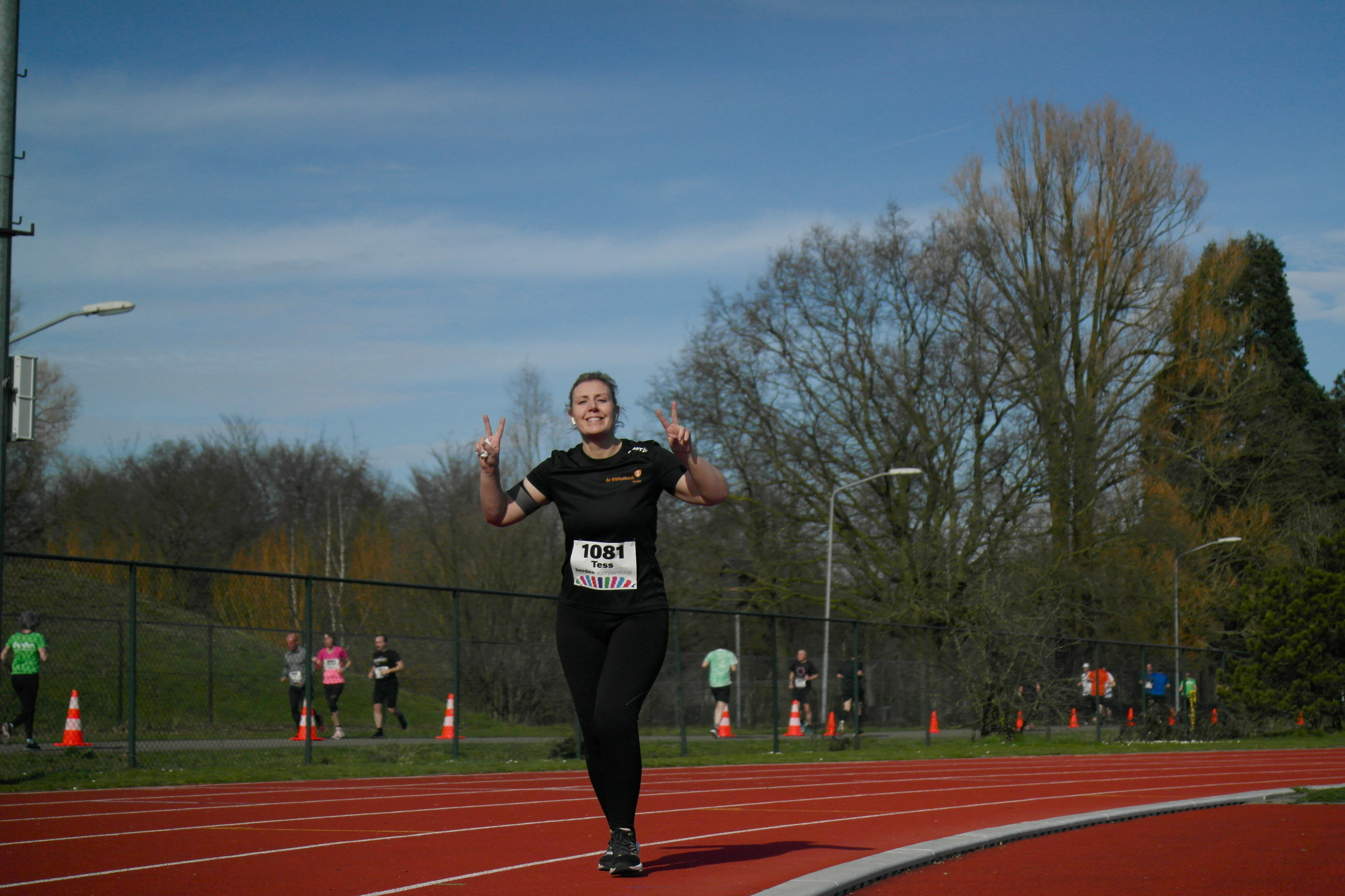 Hardloopster met startnummer 1081 maakt een vreugdegebaar tijdens de Berden Voorjaarsloop op een atletiekbaan onder een blauwe hemel.