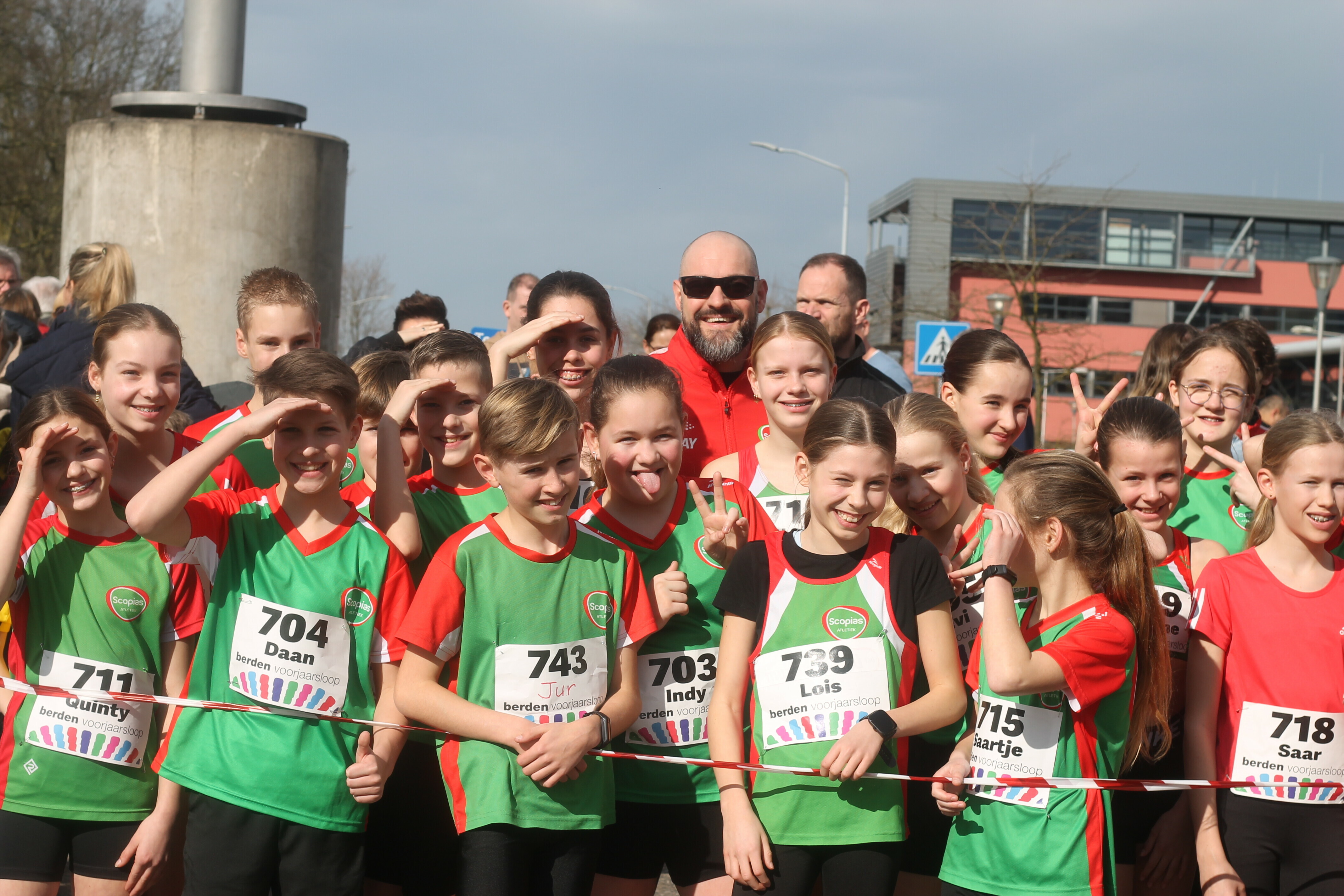 Groep jonge lopers in kleurrijke sportkleding poseert vrolijk aan de start van de Berden Voorjaarsloop, met enthousiaste gezichten en speelse gebaren.